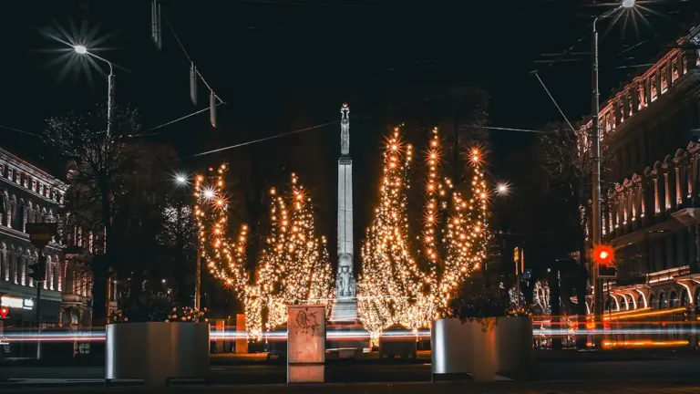 Brīvības piemineklis naktī izgaismotā Rīgas ielā ar dekoratīvām gaismiņām kokos.