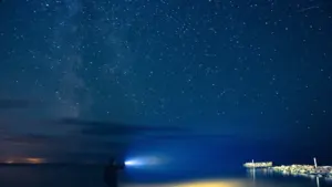 Vīrietis ar lukturīti stāv uz piestātnes pie jūras zem plašām zvaigžņotām debesīm Latvijā.