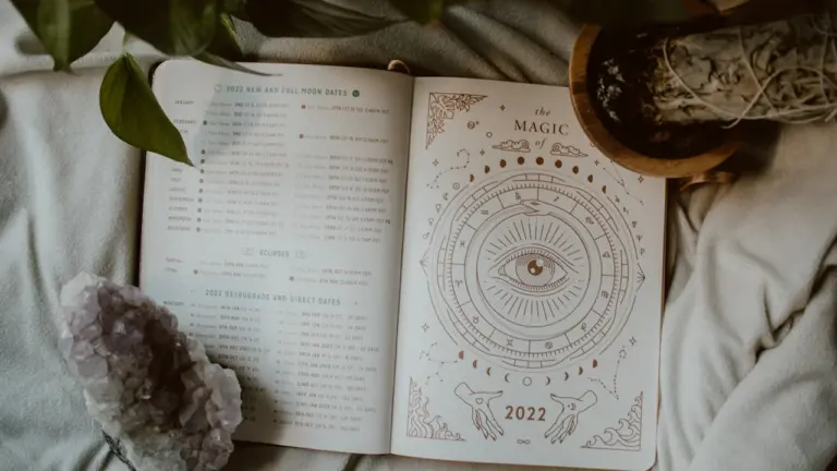 Atvērta dienasgrāmata ar astroloģiskiem simboliem, kristālu un augu uz mīksta auduma.