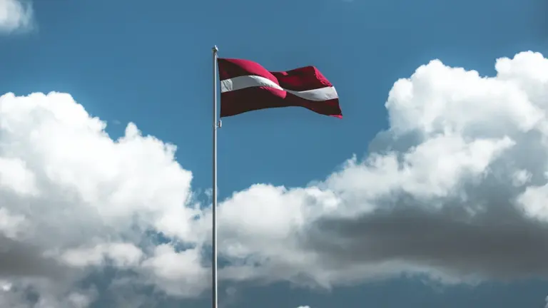 Latvijas sarkanbaltsarkanais valsts karogs plīvo vējā uz zilu debesu un mākoņu fona.