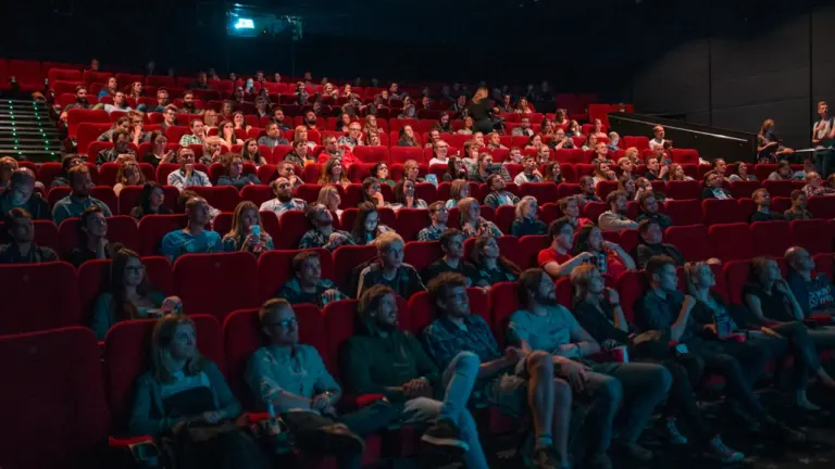 Skatītāji sēž sarkanos krēslos tumšā teātra vai kino zālē pirms izrādes sākuma.