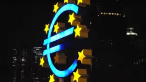 Eiropas Centrālās bankas simbols naktī, izgaismots ar zilām un dzeltenām gaismām.