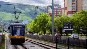 Dzelteni melns tramvajs brauc pa sliedēm Rīgas pilsētvides noskaņai atbilstošā pilsētas ainavā.
